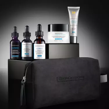 Eine Kollektion von Skinceuticals-Produkten, angeordnet auf einem schwarz-grauen, abgestuften Display vor einem dunklen Hintergrund mit Farbverlauf. Ein CE Ferulic, ein P-TIOX, ein HA Intensifier Multi-Glycan, ein A.G.E. Interrupter Advanced und ein Ultra Facial UV Defense Sunscreen. Im Vordergrund die schwarze Skinceuticals-Tasche.