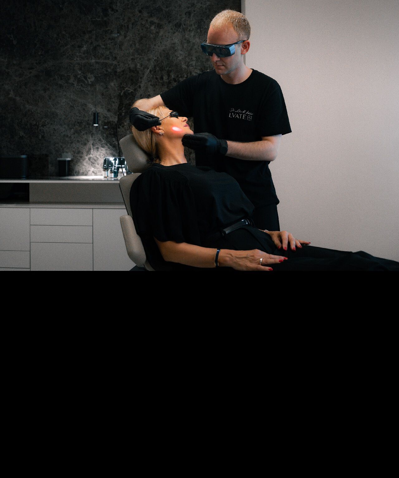 Ein Arzt in Schutzkleidung und Schutzbrille fuhrt eine Laserbehandlung im Gesicht einer liegenden Patientin durch.​
