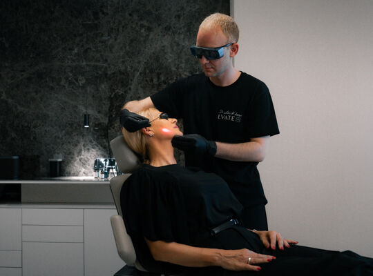 Ein Arzt in Schutzkleidung und Schutzbrille fuhrt eine Laserbehandlung im Gesicht einer liegenden Patientin durch.​