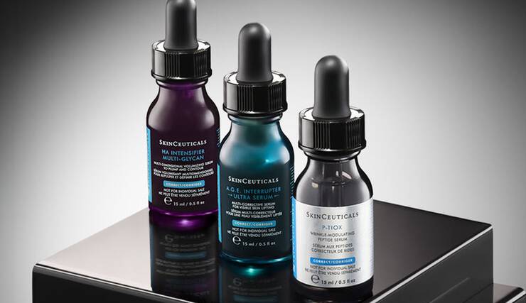 Eine Nahaufnahme von drei SkinCeuticals Hautpflegeprodukten: einem C E Ferulic Serum, einer A.G.E. Interrupter Advanced Creme und einem Advanced Brightening UV Defense Sunscreen SPF 50, die sich alle auf einer dunklen Oberflache spiegeln.