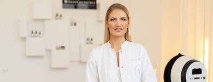 Eine lachelnde Frau in einem weißen Kittel steht in einem modernen Behandlungsraum mit einer hellen Wand und Produkten im Hintergrund.