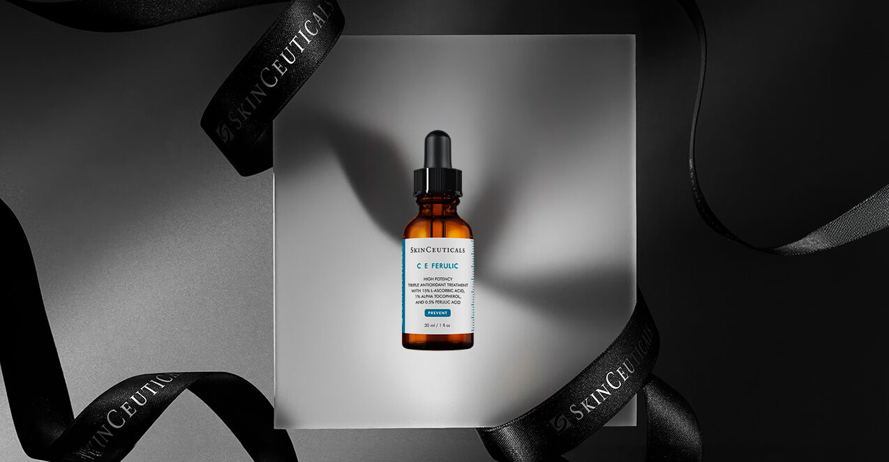 A brown glass serum bottle labeled "C E Ferulic" is displayed on a gray pedestal, surrounded by elegant black ribbons featuring the "SkinCeuticals" brand name.