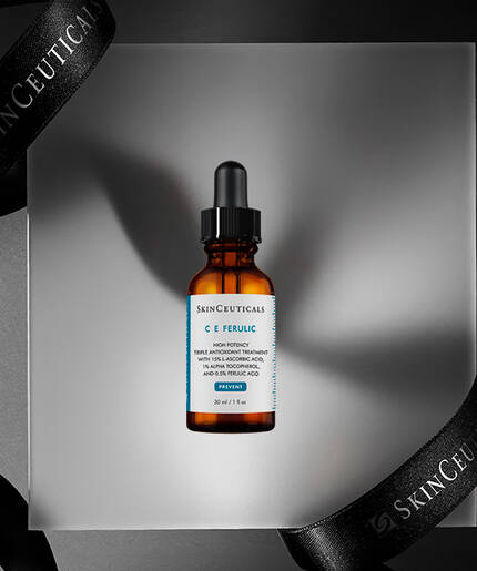 A brown glass serum bottle labeled "C E Ferulic" is displayed on a gray pedestal, surrounded by elegant black ribbons featuring the "SkinCeuticals" brand name.