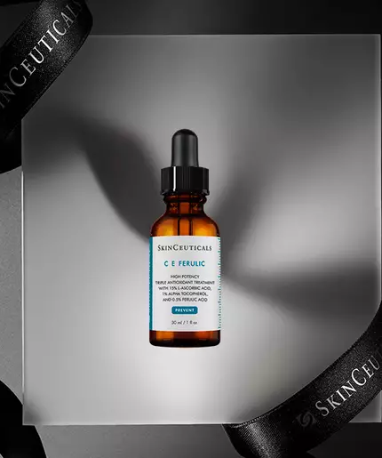 A brown glass serum bottle labeled "C E Ferulic" is displayed on a gray pedestal, surrounded by elegant black ribbons featuring the "SkinCeuticals" brand name.