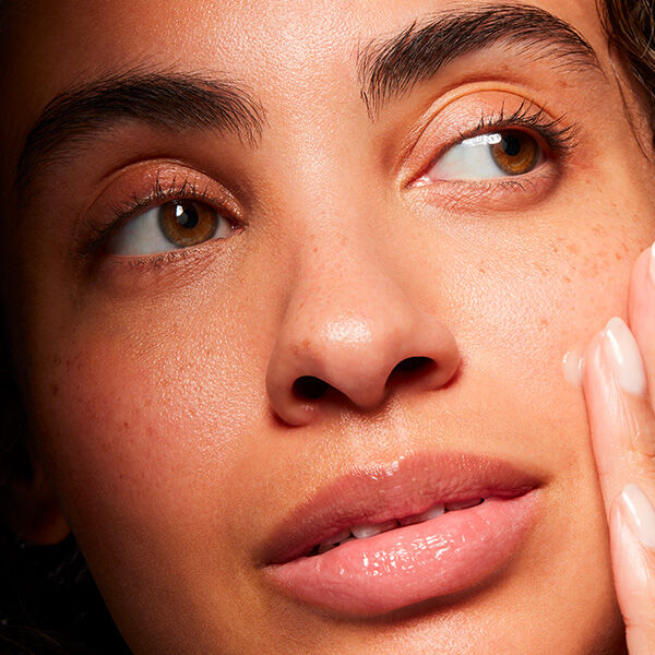 A close-up of a person's face, featuring smooth skin, brown eyes, and subtle freckles. The individual is gently touching their cheek, with a soft expression and natural makeup.