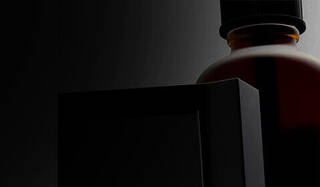 A close-up of a dark bottle and a stylized black object, set against a light gray background. The focus is on the unique shapes and textures.