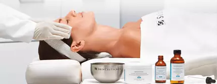 Eine Frau liegt entspannt auf einer Liege, wahrend eine Fachkraft ihre Gesichtshaut behandelt. Neben ihr stehen Produkte von SkinCeuticals auf einem Tisch.