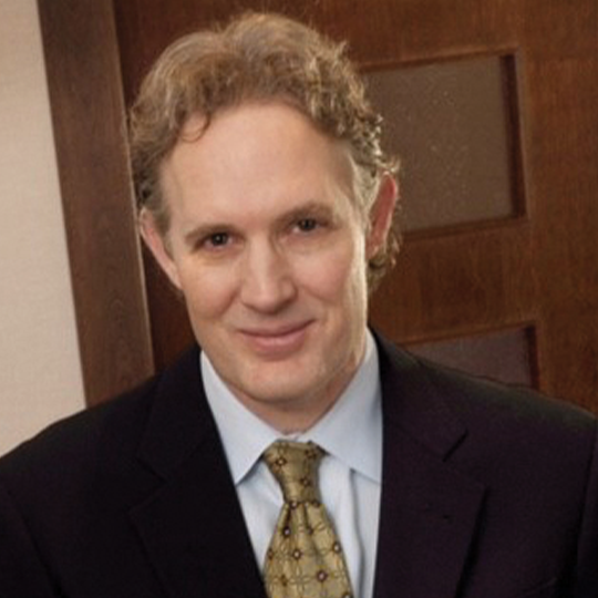 A smiling man with curly hair wearing a dark suit and tie, standing in front of a wooden-paneled background.